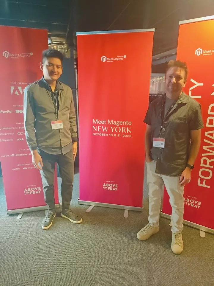 Two Towering Media team members at Meet Magento New York 2023, standing in front of the event banners